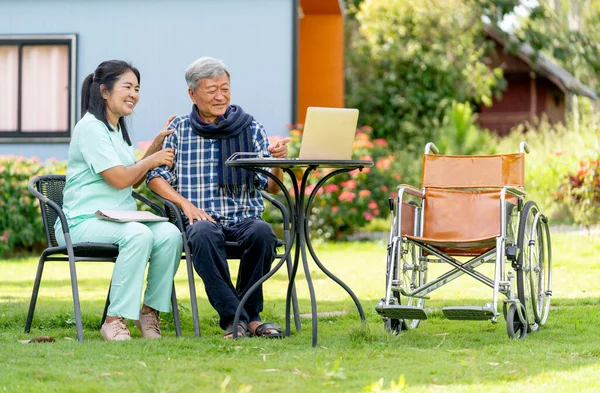 Asian senior man and nurse enjoy to watch the entertainment from laptop in area of garden with warm light.