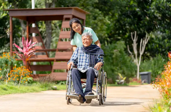 Asian nurse cart of shove senior man sit on wheelchair to walk along the walkway in the garden and they look happiness outdoor to relax with warm light.