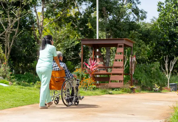 Back of Asian nurse cart of shove senior man sit on wheelchair to walk along the walkway in the garden and they look happiness outdoor to relax with warm light.