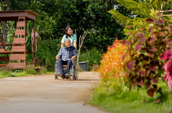 Wide shot of Asian nurse cart of shove senior man sit on wheelchair to walk along the walkway in the garden and they look happiness outdoor to relax with warm light.