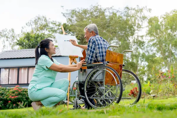 Asian nurse sit to take care near senior man sit on wheelchair and draw or paint for relax and revive good health of old people in outdoor with warm light.