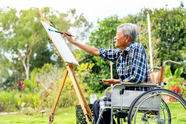 Side view of one Asian senior man sit on wheelchair and paint the painting outdoor for relax and revive good health of old people outside.