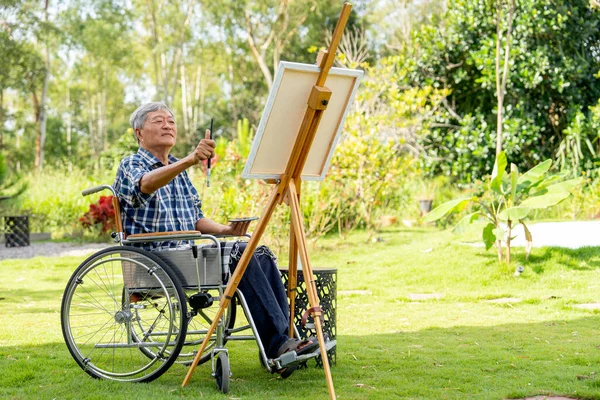 Asian senior man sit on wheelchair and action of aim or point to the area of model for paint the painting outdoor for relax outside.