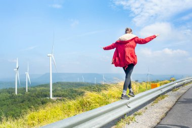 Kırmızı paltolu bir kadın rüzgar türbinlerinin ya da dağdaki yel değirmenlerinin yakınındaki bariyer çitinde yürüyor..