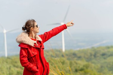 Kırmızı ceketli beyaz kadın gülümsüyor ve rüzgar türbinlerinin ya da dağdaki yel değirmenlerinin önünde sağa doğru işaret ediyor..