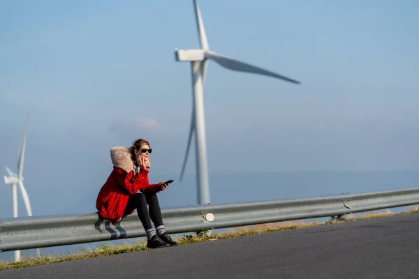 Caucasian woman with red coat and sunglasses sit on barrier of roadside and use mobile phone on mountain near windmill or wind turbine with morning light.