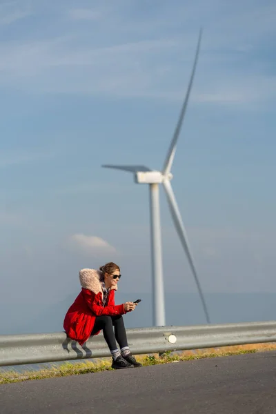 Vertical image of Caucasian woman with red coat and sunglasses sit on barrier of roadside and use mobile phone on mountain near big windmill or wind turbine.