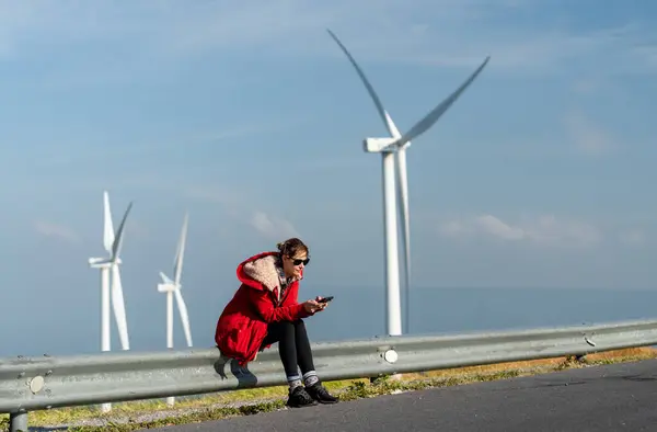 Caucasian woman with red coat and sunglasses sit on barrier of roadside and use mobile phone on mountain near windmill or wind turbine with morning light.