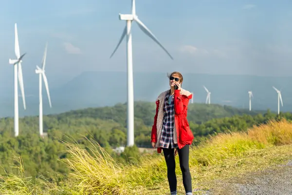 Wide shot of Caucasian woman with red coat and sunglasses stand near the roadside and use mobile phone on mountain near windmill or wind turbine with morning light.