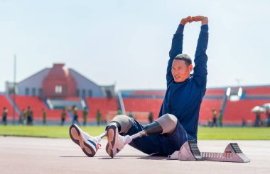 Spor adamı atleti, spor konsepti olan stadyumda oturarak ve ellerini başının üstüne koyarak egzersizden önce vücudunu ısıtmak için yaptığı bacak hareketi üzerine protez takıyor..