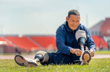Ön manzara ve bacağında protezi olan sporcu adam stadyumun yanındaki piste ayakkabı bağcığı bağlamak için çim tarlasına oturur. Spor konsepti.