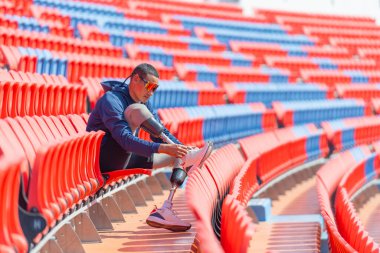Bacağında protezi olan sporcu bir sporcu stadyumdaki ayakkabı bağcıklarını gün ışığında bağlamak için amfi tiyatronun sandalyesine oturur..