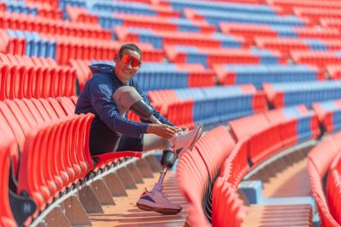 Bacağında protez olan sporcu bir sporcu ayakkabı bağcığı bağlamak için amfi tiyatronun sandalyesine oturur ayrıca gülümser ve gün ışığında stadyumdaki kameraya bakar..