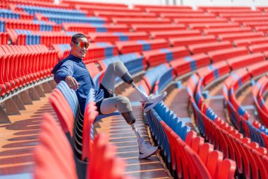 Sporcu atlet protez bacakları, amfi tiyatronun sandalyesine oturur bacaklarına poziton verip, gün ışığında stadyumdaki kameraya bakar..