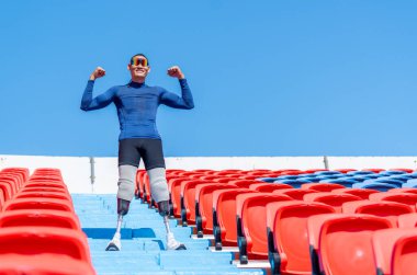 Spor adamı protez bacaklarının yan görünüşü mutlu bir şekilde merdivenlerde duruyor ve gün ışığında stadyumdaki amfitiyatroda kutlama yapıyor..