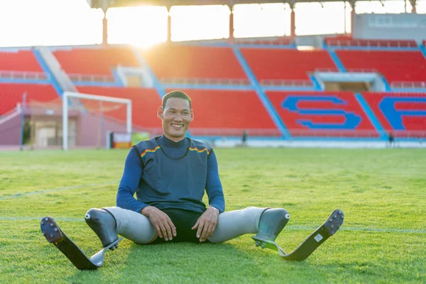 Sport man athlete prosthesis legs sit down on grass field and separate his legs to relax also look at camera with smiling in the stadium.