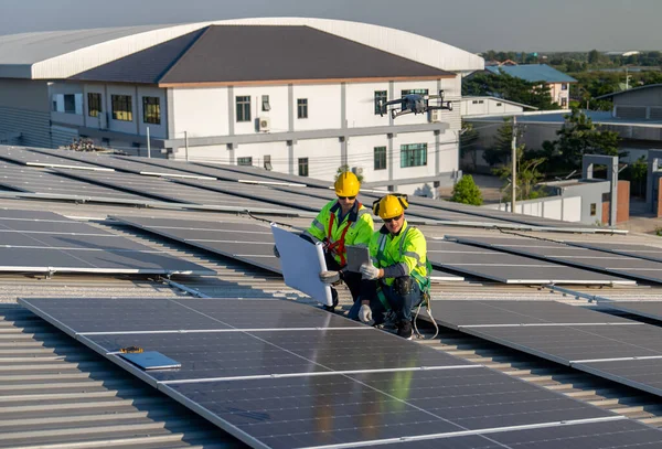 Side view of two Caucasian technician workers use construction plan and tablet to check and maintenance solar cell panels on rooftop of factory or the building. Green and sustainable energy concept.