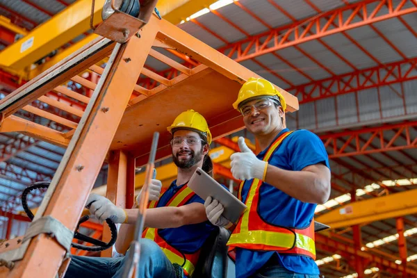 Two Caucasian factory worker show thumbs up and look at camera with one sit in truck and other stand beside with smiling in factory workplace area.