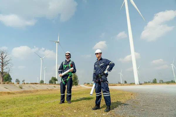 Professional Caucasian workers or technician with safety suit hold construction paper plan and stand in front of wind turbine or windmill in the row as power plant business.