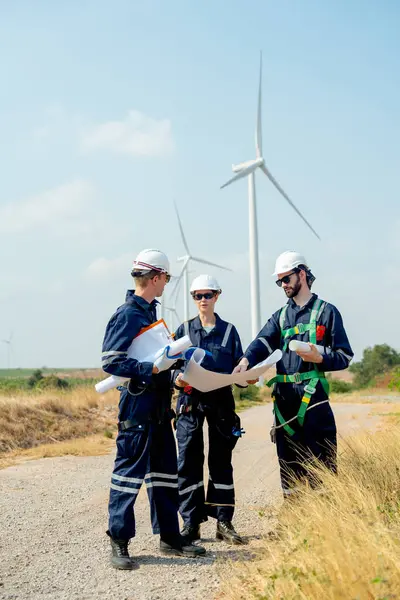 Vertical image of professional technician man and woman workers discuss about work project on plan paper in front of wind turbine or windmill in workplace area.