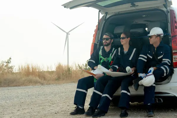 Close up group of technician workers sit on back of van and discuss about work in plan paper on the road with windmill or wind turbine on the background.