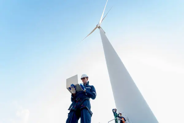 Professional technician man stand with hold laptop and stand near base of windmill or wind turbine with his co-worker in the back.