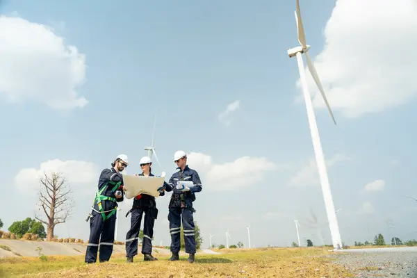 Group of professional technician workers stand with discuss the project in the paper plan and stand in front of wind turbine or windmill in area for power plant business.