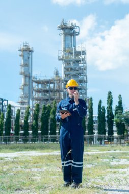 Fabrika mühendisi ya da teknisyeninin dikey görüntüsü elinde telsiz olan ve gün ışığında rafineri ve petrokimya fabrikasının önünde duran.