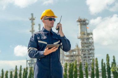 Fabrika mühendisi ya da teknisyen telsizi elinde tutarken tableti de elinde tutuyor ve gün ışığında rafine ve petrokimya fabrikasının önünde duruyor..