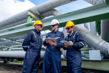 Mühendis ve fabrika teknisyenlerinden oluşan ön ve alt görüş grubu petrokimyasal gaz boru hattının yanında duruyor ve çalışmalarını tartışmak için tablet kullanıyorlar..