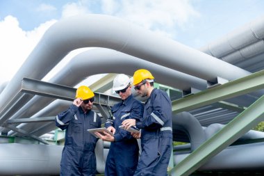 Mühendis ya da fabrika teknisyenlerinden oluşan bir grup petrokimyasal gaz boru hattının yanında ve altında duruyor ve işyerlerinde yaptıkları işleri tartışmak için tablet kullanıyorlar..