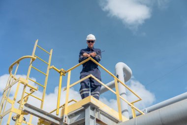 Fabrika mühendisi veya teknisyeni petrokimyasal gaz boru hattı üzerinde yüksek bir eyalette duruyor ve mavi gökyüzü arka plan olarak işyerindeki daha düşük görüntüye bakıyor.