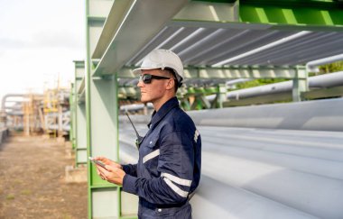 Petrokimya boru hattına dayanan mühendis veya teknisyen işçinin geniş ve yandan görünüşü ve cep telefonuyla keyfini çıkarması.
