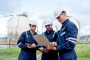Bir grup mühendis ya da teknisyen, bulutlu gökyüzü olan petrokimya fabrikasındaki tankın önünde durup dizüstü bilgisayara bakıyorlar..