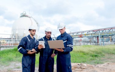 Bir grup mühendis ya da teknisyen, bulutlu gökyüzü olan petrokimya fabrikasının önünde dizüstü bilgisayara bakıyorlar..