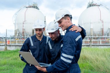 Mühendis veya teknisyen işçiler dizüstü bilgisayara bakar ve bulutlu gökyüzü ile petrokimya fabrikasının önünde dururlar..