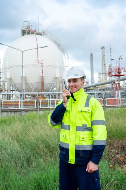 Mühendis ya da teknisyenin dikey görüntüsü gündüz ışığında petrokimya tankının önünde telsiz ile ayakta duruyor..