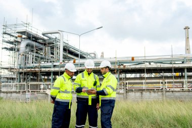 Fabrika mühendisi ya da teknisyenlerinden oluşan geniş bir grup bir tablet ve diğerini işaret ediyor ve aynı zamanda gün ışığında rafine ve petrokimya fabrikası ve tankın önünde duruyorlar..