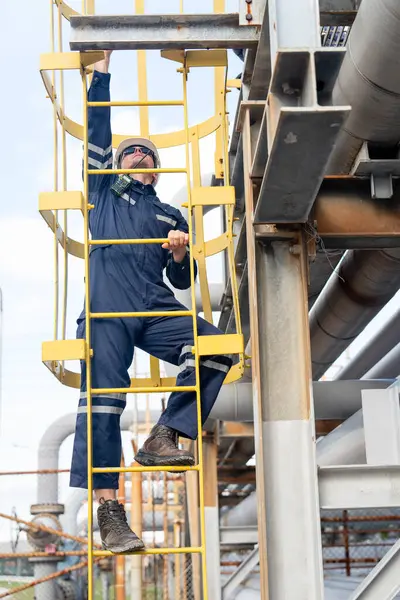 Fabrika mühendisi veya teknisyeninin dikey görüntüsü çalışmak veya işyerinde bakım yapmak için petrokimyasal gaz boru hattının merdivenlerine tırmanıyor.
