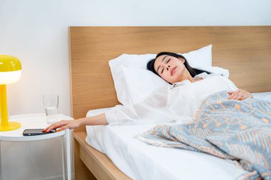 Asyalı bir kadın sabah uyandığında yatağında uyur ve yatağın yanındaki cep telefonuna dokunur..