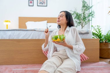 Güzel Asyalı kadın elinde bir kase sebze salatası tutuyor ve yemekten mutlu olduğunu ifade ediyor, ayrıca dinlenmek için yatak odasına yakın oturuyor..