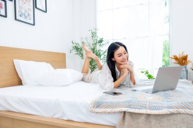 Güzel Asyalı kadının yan görünüşü yatağa uzanır ve dizüstü bilgisayarını mutlulukla kullanmaktan zevk alır..