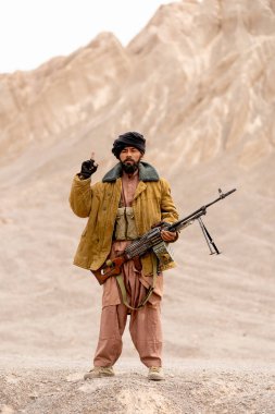 Kostümlü adam terörist gibi görünüyor. Silahı var ve dağın önünde duruyor. Kameraya bakıp gökyüzünü gösteriyor..