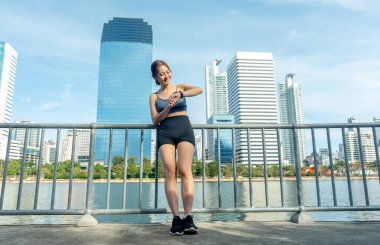 Ön manzara Asyalı kadın tırabzana eğiliyor ve kamuya açık alanda egzersiz yaptıktan sonra arka planda büyük şehir ve yumuşak ışıkla rahatlamak için saatine bakıyor..