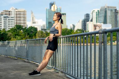 Asyalı bir kadın duvarlara yaslanmış, arka planda büyük bir şehir ve yumuşak bir ışıkla halka açık alanda egzersiz yaptıktan sonra rahatlamak için saatine bakın..