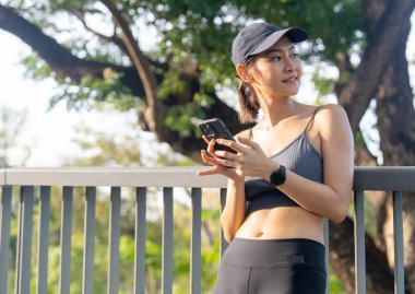 Asyalı spor kadının portresi kaldırım korkuluklarında eğiliyor ve sağa bakıyor ve halka açık bahçede egzersiz yaptıktan sonra cep telefonu kullanıyor..