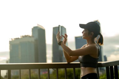 Asyalı sporcu kadın fotoğraf çekmek için cep telefonu kullanıyor ve arka planda büyük şehir ve yumuşak ılık ışık ile halka açık alanda yapılan egzersizden sonra dinleniyor..