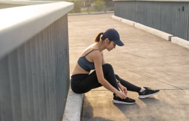 Asyalı bir kadının oturma ve yürüme köprüsündeki çitlere doğru eğilme görüntüsü de halka açık alanda akşamları sıcak ışık ve arka planda yüksek bina eşliğinde yapılan egzersizden sonra gevşemek için ayakkabı bağcıkları bağlıyor..