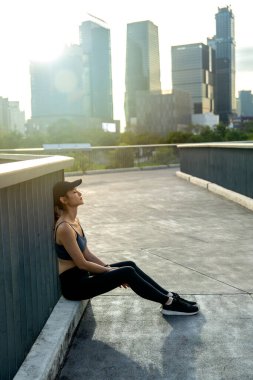 Asyalı bir kadının dikey görüntüsü yürüyüş köprüsündeki çitlere yaslanır ve ileriye bakar akşam vakti halka açık alanda egzersizden sonra dinlenir ve arka planda yüksek binalar inşa edilir..