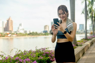 Asyalı sporcu kadın cep telefonu kullanıyor ve arka plan olarak yüksek bina ve yumuşak ışık ile kamu bahçesinde egzersiz yaptıktan sonra dinleniyor.